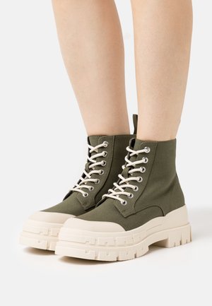 Green canvas ankle boots with beige rubber sole, lace-up design, and textured accents on the sole. Mid-top style.
