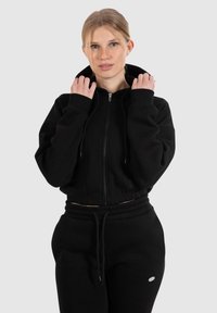 Sudadera negra corta con cremallera completa, capucha con cordón y puños acanalados, combinada con pantalones deportivos negros de cintura alta a juego.