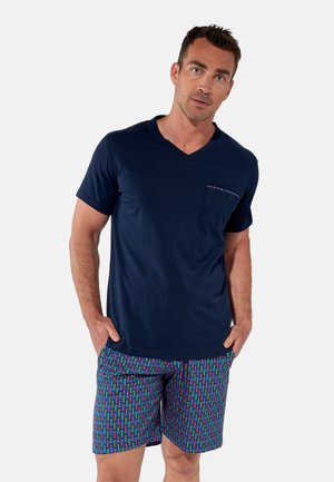 Mężczyzna w granatowej koszulce z dekoltem w serek z przednią kieszenią i wielokolorowych szortach w geometryczny wzór, stoi z rękami w kieszeniach.