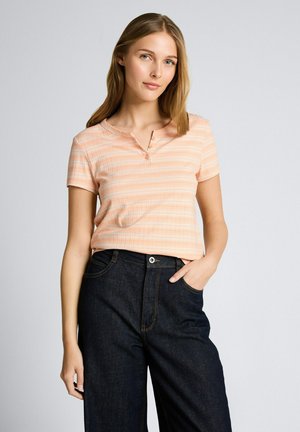 Jeune femme aux cheveux châtain clair portant une chemise à carreaux pêche à manches courtes et un jean bleu foncé taille haute, debout avec une main dans la poche.