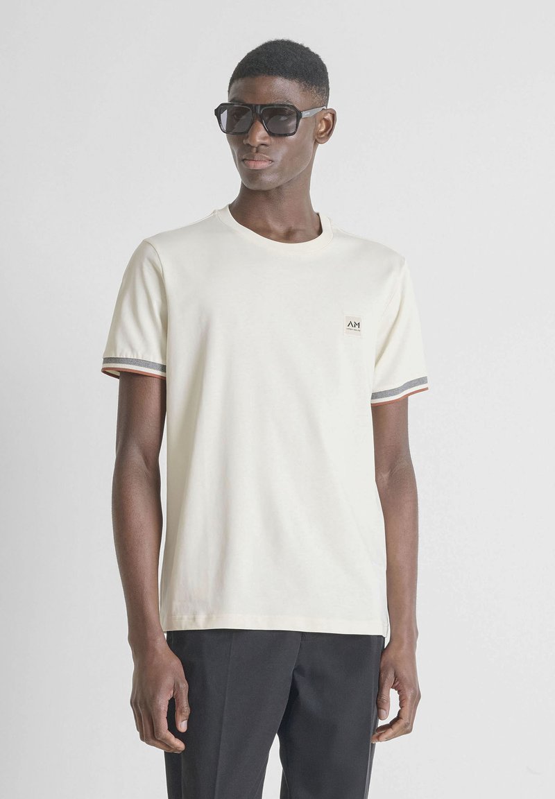 Hombre con gafas de sol negras, camisa blanca de manga corta con puños a rayas y pequeño logo, y pantalones negros contra un fondo liso.