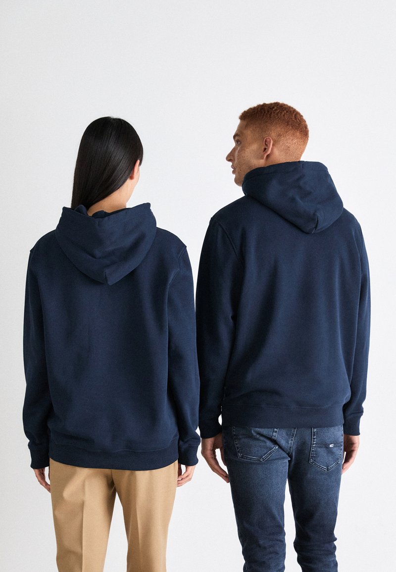 Des sweats à capuche bleu marine avec poches kangourou, poignets et coutures côtelés, associés à un pantalon beige et un jean foncé, vus de derrière.