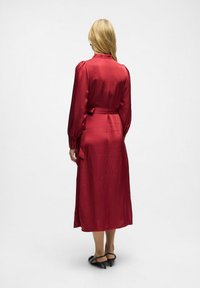 Robe en satin rouge avec des manches longues, un col volé et une taille ceinturée. Longueur mi-mollet avec une texture lisse et une silhouette fluide.