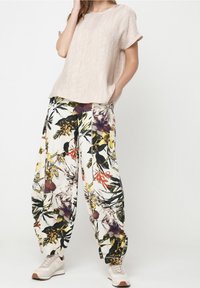 Vrouw in een beige, gestructureerd T-shirt met korte mouwen, wijde bloemenprint broek en beige sneakers, staand met één hand in de zak.