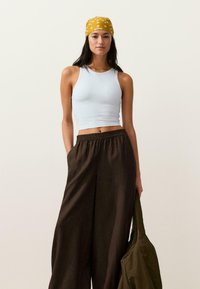 Frau mit gelbem, gemustertem Kopftuch, weißem ärmellosem Crop-Top, weitem braunen Hosen und einer olivgrünen Jacke an der Seite haltend.