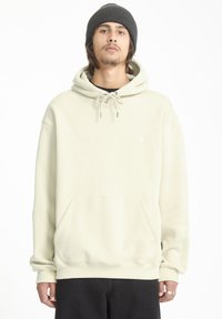 Lichtbeige hoodie met een kangoeroezak, trekkoordcapuchon, geribbelde manchetten en een klein logo op de borst, gecombineerd met zwarte broek.