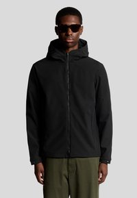 Veste à capuche noire avec fermeture éclair, présentant une texture lisse et un design pratique. Portée sur un pantalon vert, avec des lunettes de soleil foncées.