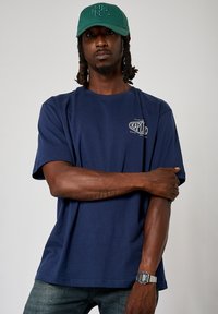 T-shirt en coton bleu marine avec des manches courtes et un logo sur le devant. Porté avec une casquette verte. La personne a une montre-bracelet en argent et un tatouage.