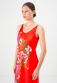 Mujer con el cabello recogido que lleva un vestido rojo sin mangas con un patrón floral multicolor en el frontal, mirando hacia un lado.