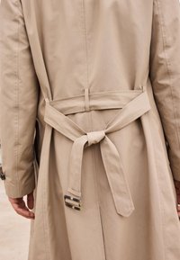 Gabardina beige con un cinturón atado en la cintura, que presenta una textura suave y una hebilla rectangular y transparente en el cinturón.