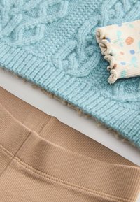 Ljusblå, texturerad stickad tröja med ribbad nederkant ovanför beige ribbade byxor. En liten mönstrad tygbit med färgglada prickar ingår.