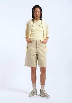 Jonge vrouw die beige korte broek draagt, lichtgele blouse en een open korte mouwen blouse met knopen, staand met handen in de zakken op een witte achtergrond.