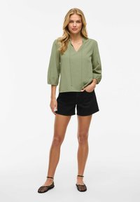 Top verde chiaro a V con cordino, abbinato a pantaloni corti neri. L'outfit presenta un fit rilassato e maniche 3/4, scarpe basse con dettagli in rete.