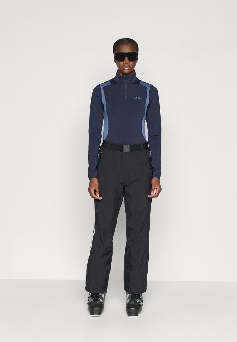 Haut à manches longues navy avec des accents bleu clair, col montant ; pantalon de ski noir avec des rayures blanches sur les côtés ; modèle portant des bottes de ski noires et des lunettes de soleil.