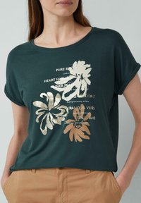 Groene t-shirt met korte mouwen van zachte stof, met gouden bloemenprint en tekst in het midden, gecombineerd met lichtgekleurde broeken.