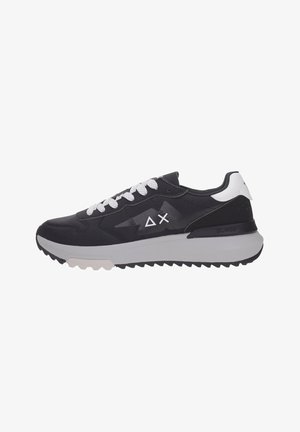 Zapatilla deportiva baja en blanco y negro con cordones blancos, suela blanca gruesa y símbolos de logotipo en el lateral y el talón.