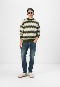Grønn og kremfarget zigzag mønstret strikkegenser med ribbet ermer, kombinert med blå jeans og hvite joggesko. Modellen har på seg runde solbriller.