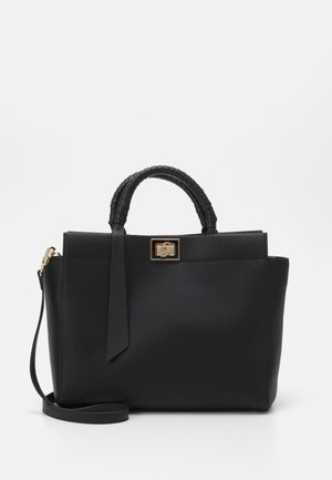 Sac à main texturé noir avec poignées courtes enveloppées, fermoir rectangulaire doré, longue bandoulière, et un ruban noir décoratif sur fond blanc.