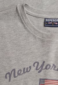 Grijze stoffen T-shirtkraag met de tekst "New York" en een label met gekruiste Britse en Amerikaanse vlaggen en het merk "SUPERDRY".