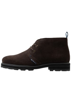 Dunkelbrauner Wildleder-Desert-Boot mit zwei Ösen, schwarzer Gummisohle und einer Zuglasche hinten, seitlich auf weißem Hintergrund dargestellt.
