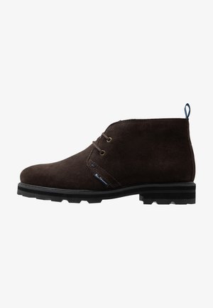 Botte désert en daim brun foncé avec deux œillets, semelle en caoutchouc noire et une languette arrière, vue de profil sur fond blanc.