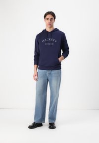 Námořnický svetr s kapucí a přední kapsou typu klokan, s logem "HACKETT LONDON". V kombinaci s světle modrými širokými džíny a černými botami.