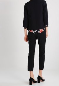 Blazer noir court porté sur un chemisier fleuri, associé à un pantalon noir ajusté et des chaussures à talons blocs noires. Tissu lisse, design structuré.