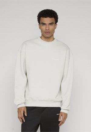 Man met kort krullend haar, gekleed in een effen wit sweatshirt met lange mouwen en een zwarte broek, staand tegen een lichtgrijze achtergrond.