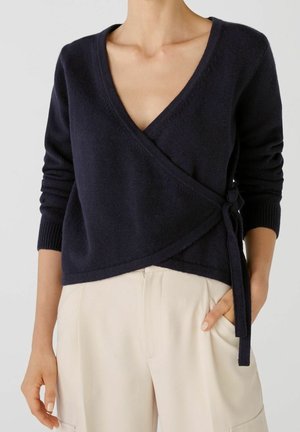 Kvinde iført en mørk navy slå-om-sweater med lange ærmer og beige bukser, hånd i lommen, mod en ensfarvet lys baggrund.
