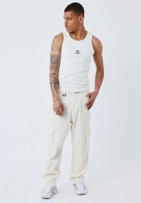 Canotta bianca con logo "PXP" nero sul petto, abbinata a pantaloni cargo crema con tasche laterali. Il modello indossa sneakers grigio chiaro.