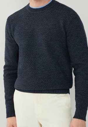 Man wearing a dark blue textured knit sweater over a light blue shirt, paired with cream-colored pants against a plain background.