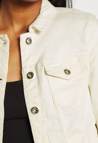 Vit denimjacka med metallknappstängning, en bröstficka och en klassisk kragsdesign med en mjuk, texturerad yta.