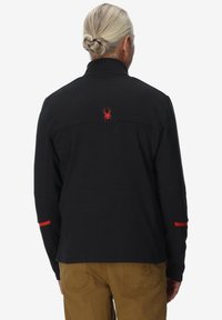 Uomo con capelli grigi raccolti in un bun, che indossa una giacca nera con un logo di ragno rosso sulla parte alta della schiena e pantaloni marroni, di spalle.