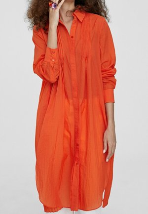 Donna che indossa un lungo abito camicia arancione brillante con pieghe vicino al colletto, che tiene la mano vicino al viso con anelli colorati.