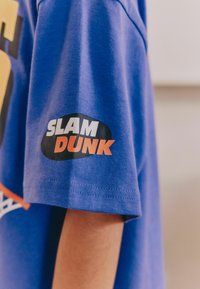 Blå bomullströja med korta ärmar som har en svart oval lapp med texten "SLAM DUNK" i vitt och orange, placerad på ärmen.