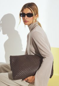 Woven braune Leder-Clutch mit strukturierten Oberflächen. Modell trägt einen beigen Rollkragenpulli und hält die Tasche vor einem hellen Hintergrund.