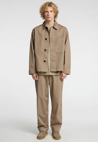 Tanfarget jakke med knappestengning og en enkelt brystlomme, kombinert med en matchende tanfarget skjorte og bukser. Semskede sko med gummisåler.