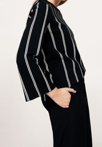 Pull rayé noir avec des lignes verticales blanches, doté de manches larges et d'une coupe décontractée. Le modèle porte le pull avec un pantalon foncé.