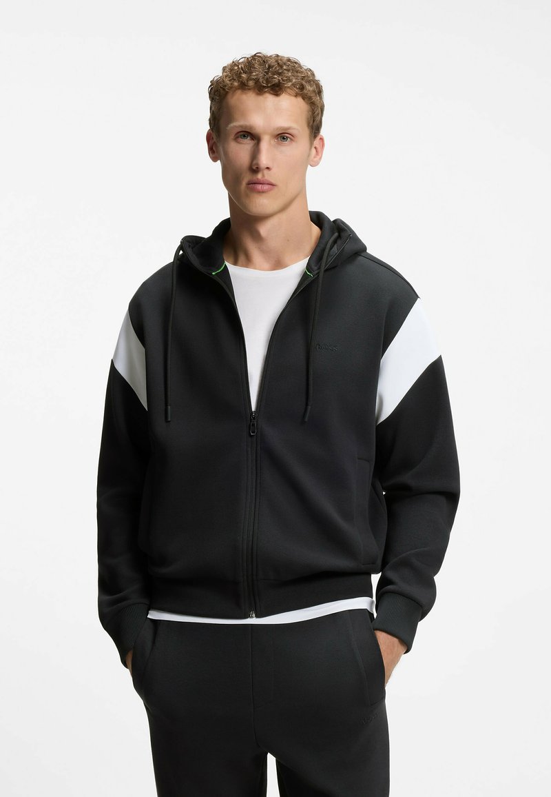 Jeune homme aux cheveux bouclés porte un sweat à capuche zippé noir avec des rayures blanches diagonales sur le haut des manches et un pantalon noir assorti.