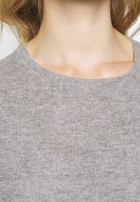 Pull en maille grise à texture lisse, avec un col rond classique et une coupe ajustée, mettant en valeur la partie supérieure de la poitrine.