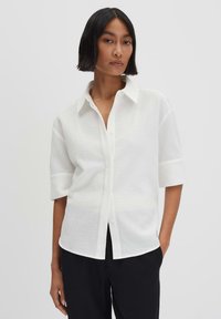 Camicia bianca a maniche corte con tessuto testurizzato, colletto classico e vestibilità comoda. Indossata con pantaloni neri.