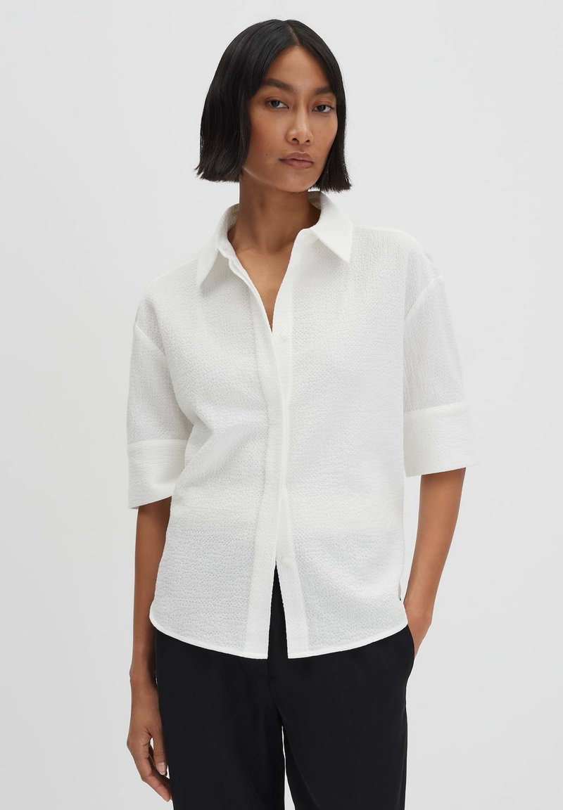 Camicia bianca a maniche corte con tessuto testurizzato, colletto classico e vestibilità comoda. Indossata con pantaloni neri.