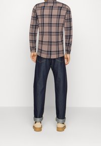 Mężczyzna w ciemnoniebieskich jeansach z podwiniętymi nogawkami, beżowej i czarnej koszuli w kratę z długim rękawem oraz beżowych butach, stojący na tle białego tła.