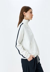 Veste zippée blanche en matière douce avec un col haut et des rayures latérales navy, comportant des poches avant et une coupe décontractée.