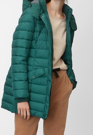 Femme portant une veste matelassée verte à capuche sur un haut beige et un pantalon marron, une main dans la poche et l'autre détendue à ses côtés.