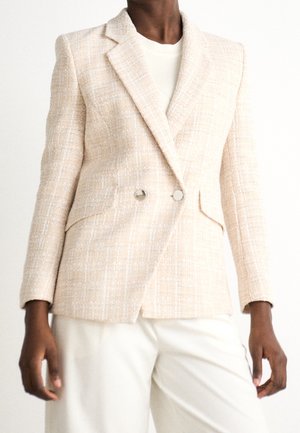 Person trägt einen cremefarbenen, strukturierten, zweireihigen Blazer über einem weißen Oberteil und weißen Hosen vor einem schlichten weißen Hintergrund.
