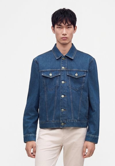 Jeune homme portant une veste en jean bleue boutonnée avec des poches poitrine, debout devant un fond clair uni.