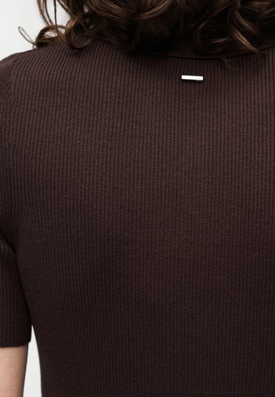 Personne aux cheveux bouclés foncés portant un pull marron côtelé à manches courtes avec une petite épingle rectangulaire argentée dans le haut du dos.