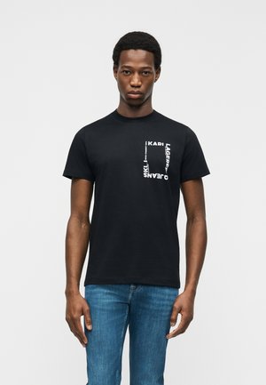 Mand iført en sort T-shirt med hvidt tekst i firkantet form på venstre bryst og blå jeans, stående mod en ensfarvet baggrund.