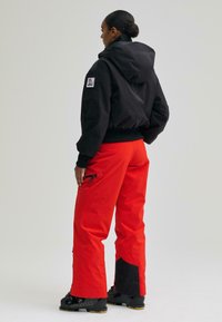 Jachetă neagră cu glugă și manșete strâmte, asociată cu pantaloni roșii strălucitori ce au buzunare cu fermoar și accente negre la tiv.